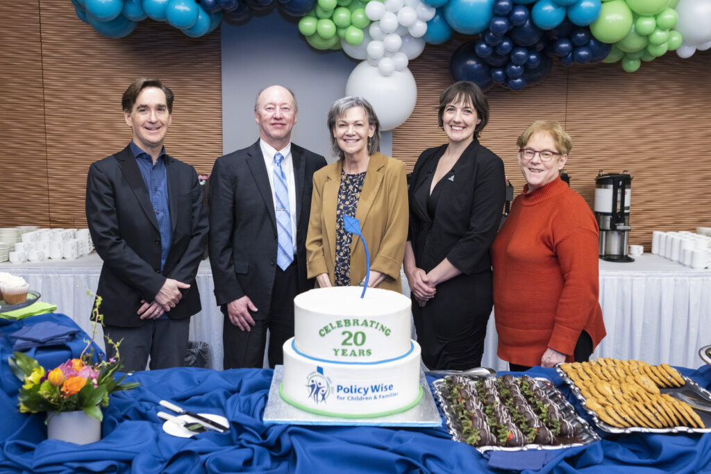 Members of PolicyWise's Board of Directors are gathered around a 20th anniversary cake.