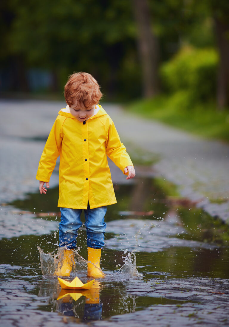 cute baby boy in yellow raincoat and rubber boots having fun, jumping in puddles, launching the paper boat after spring rain - Generating and sharing knowledge