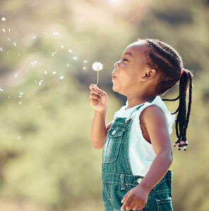 Happy little african american girl blowing a flower in outside. Cheerful child having fun playing and blowing a dandelion into the air in a park. Kid having fun with joy playing with a plant outdoors. - Strengthening Capacity