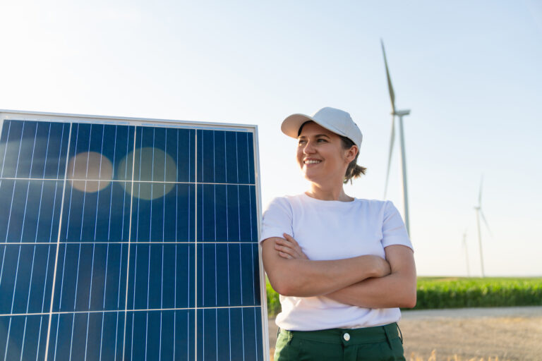 Woman wearing white cap stands next to solar panel. Wind turbines in the background.