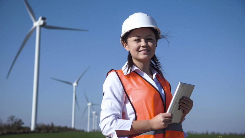 Cheerful woman wearing hard hat standing against turbines at wind farm on sunny day. Image for Opportunities for Women in Alberta’s Green Energy Sector