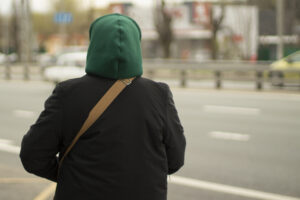 An unknown young person on the road, warmly dressed in a green hood and black jacket. The image is used for PolicyWise for Children & Families' report that research points to childhood roots of youth homelessness.