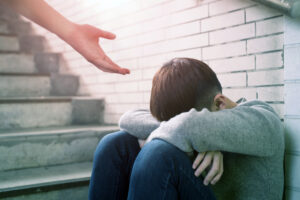 A young man who is homeless sit in an underground stairwell. A hand is reaching to provide them help. This is image is being used for PolicyWise's news story "New research shows how youth-centred information practices can help prevent youth homelessness"
