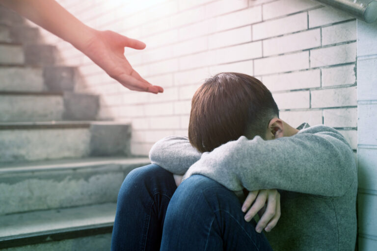 A young man who is homeless sit in an underground stairwell. A hand is reaching to provide them help. This is image is being used for PolicyWise's news story "New research shows how youth-centred information practices can help prevent youth homelessness"