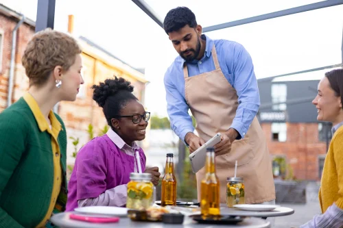 At a restaurant, a server is helping a guest pick an item from the menu. Image for PolicyWise's news story titled. Building inclusive workplaces across Alberta’s food and beverage sector