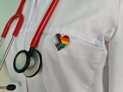 A doctor wearing a pride progress pin. This image is being used for PolicyWise's Empowering Providers Serving Gender-Diverse Youth Initiative and the supporting gender-diverse youth webinar.