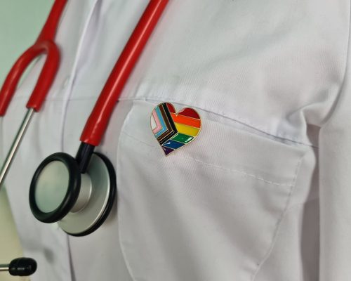 A doctor wearing a pride progress pin. This image is being used for PolicyWise's Empowering Providers Serving Gender-Diverse Youth Initiative and the supporting gender-diverse youth webinar.